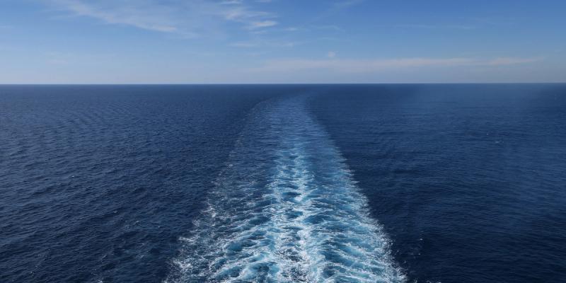 Open blue ocean with a long white wake trailing from a ship under a clear sky, evoking the global trade journey.