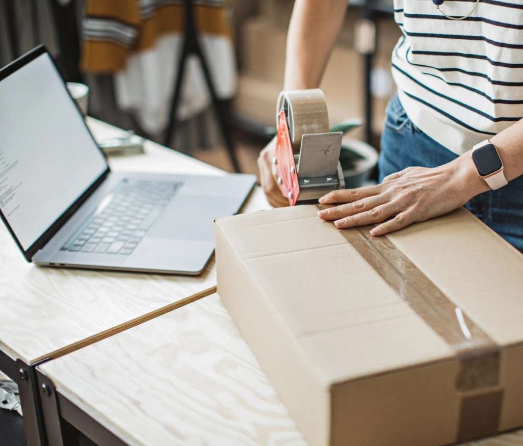 Person taping a cardboard shipping box beside an open laptop on a wooden table