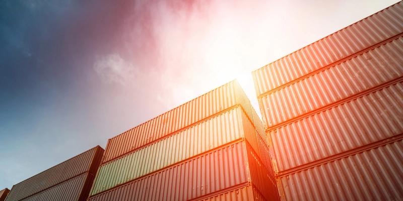 Stacks of shipping containers viewed from below with sun flare between them and a partly cloudy sky