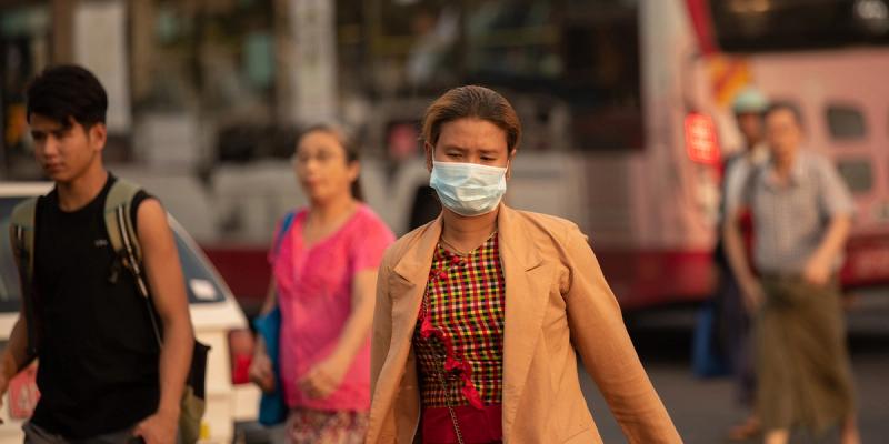 Woman in a tan blazer wearing a surgical face mask walks down a city street as pedestrians and a bus pass behind her.