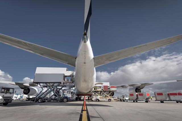 Rear view of a wide-body cargo jet on the tarmac being loaded by scissor-lift loaders, ground trucks and ULD pallets under a blue sky