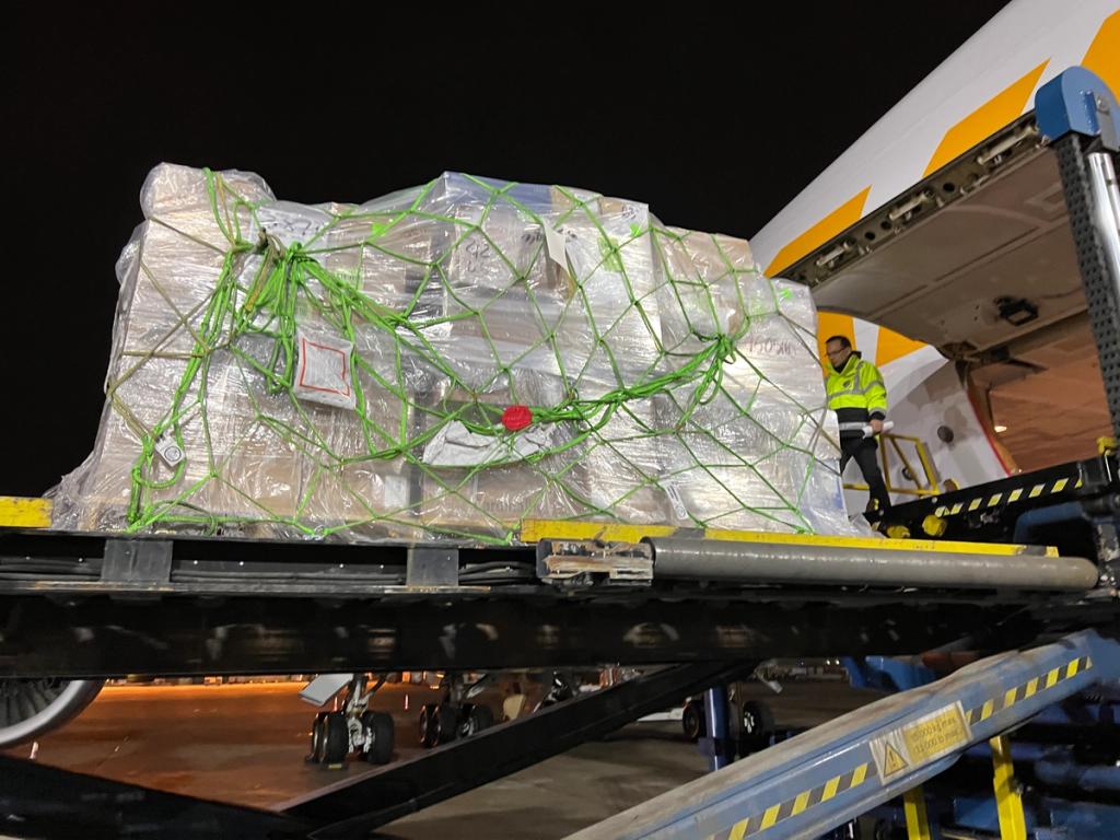 Worker in hi-vis vest loads a pallet of shrink-wrapped cargo netted in green straps onto an aircraft at night.