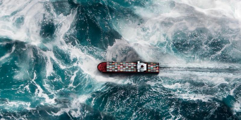 Aerial view of a container ship cutting through churning turquoise ocean waves.