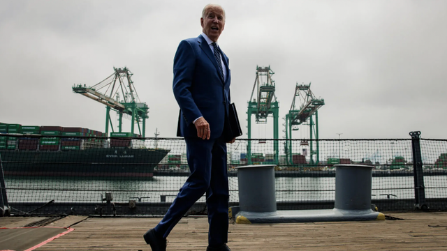 Man in a dark blue suit walking along a pier with shipping port cranes and a docked container vessel behind him