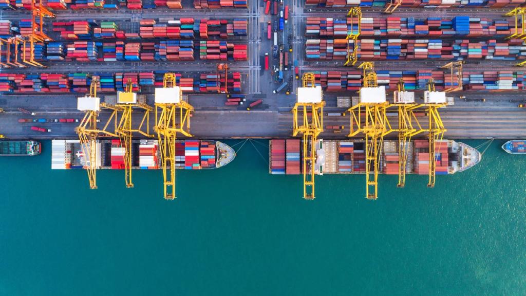Aerial view of a container terminal with yellow gantry cranes loading two ships beside stacked containers and turquoise water