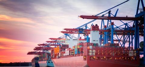A container ship docked at a port terminal at sunset, loaded with stacked containers beneath blue and red gantry cranes under a pink and orange sky.