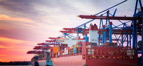 A container ship docked at a port terminal at sunset, loaded with stacked containers beneath blue and red gantry cranes under a pink and orange sky.