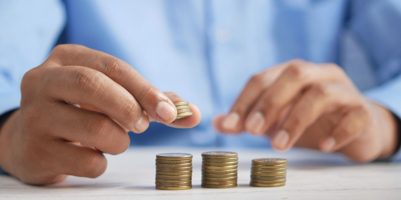 Person in a blue shirt stacking coins into rising piles on a table, suggesting cash flow and savings