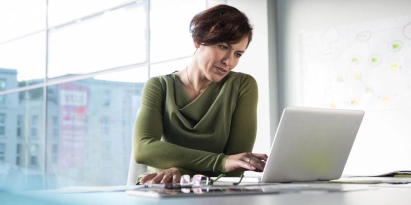 Woman in a green top working on a laptop at a desk by a window, eyeglasses resting nearby