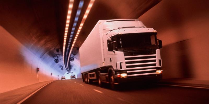 White cargo truck with trailer speeding through a lit highway tunnel, cars approaching from behind