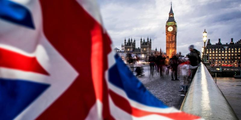 A British Union Jack flag in foreground with Big Ben and the Houses of Parliament lit at dusk across the Thames.