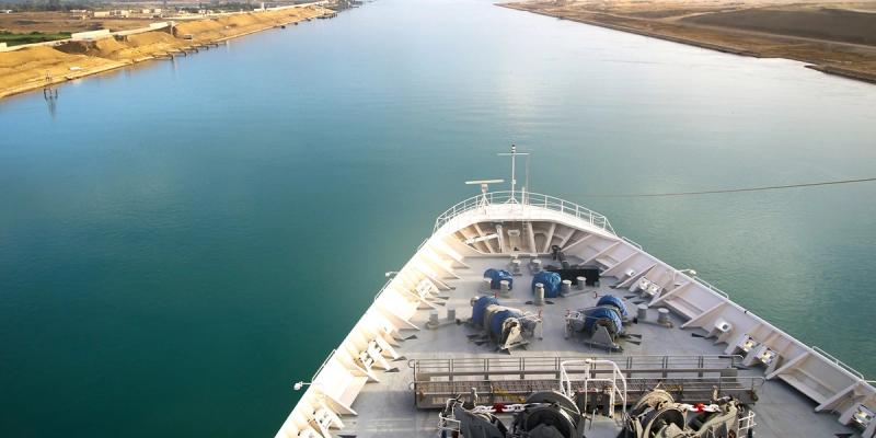 View from the bow of a large ship transiting a narrow canal with sandy banks on either side
