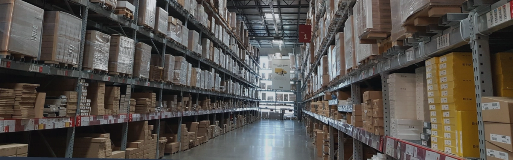 Long warehouse aisle flanked by tall industrial racks packed with boxed inventory receding to the back wall.