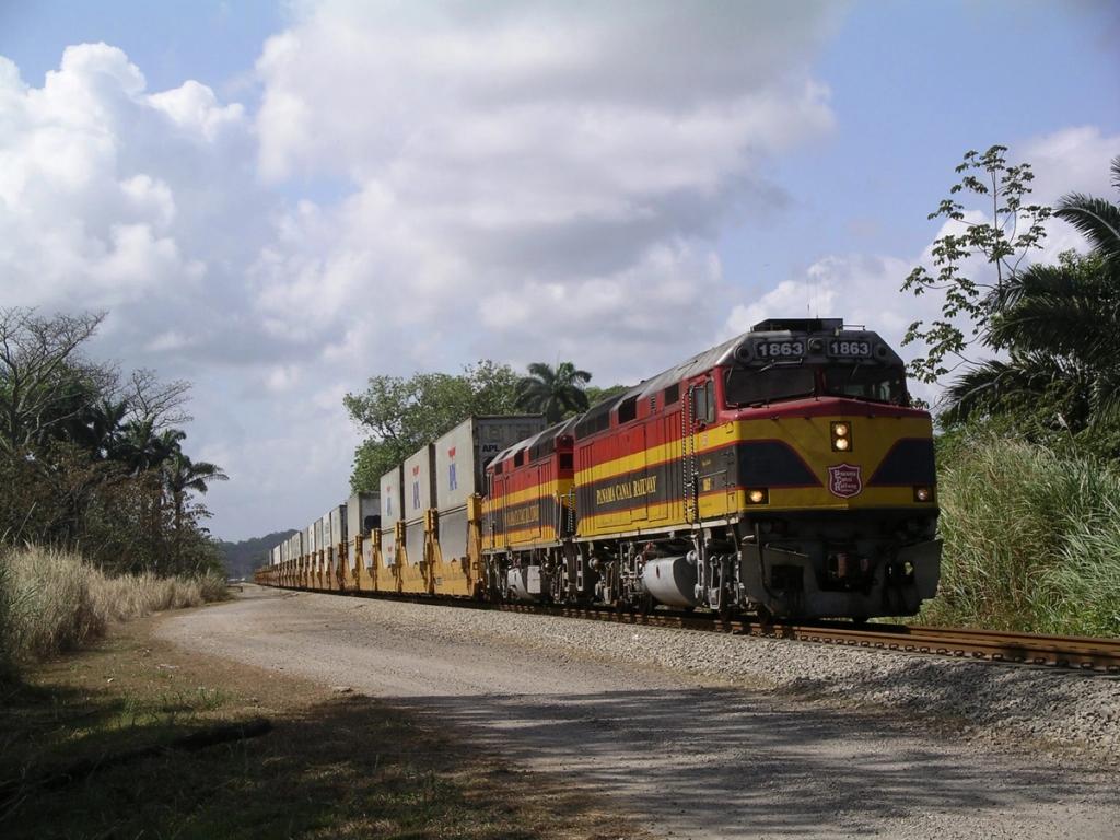 Red and yellow Panama Canal Railway locomotives haul a container train past a rural roadside.
