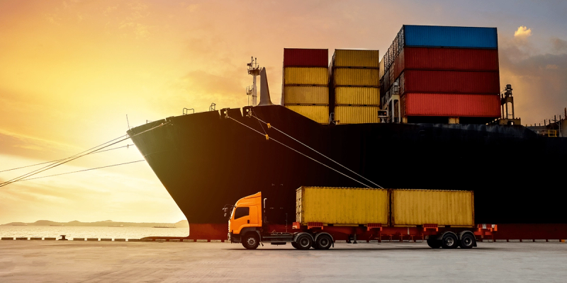Orange truck with a container beside a large container ship at a port during sunset