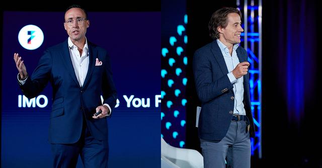Two men in suits speak on stage at a Flexport event with blue lighting and stage trusses.
