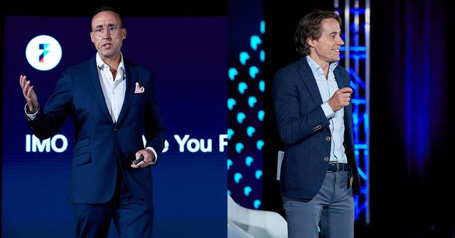 Two men in suits speak on stage at a Flexport event with blue lighting and stage trusses.