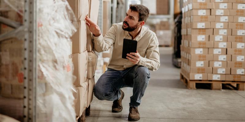 Bearded man in a warehouse aisle crouching to inspect cartons while holding a tablet