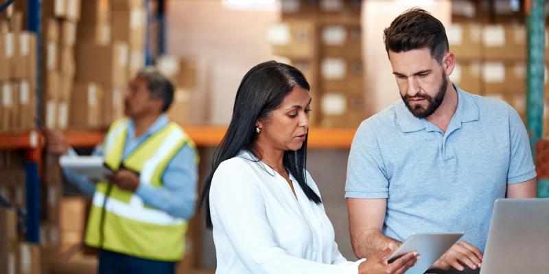A woman and man review a tablet and laptop together in a warehouse aisle as a worker in a hi-vis vest checks stock behind