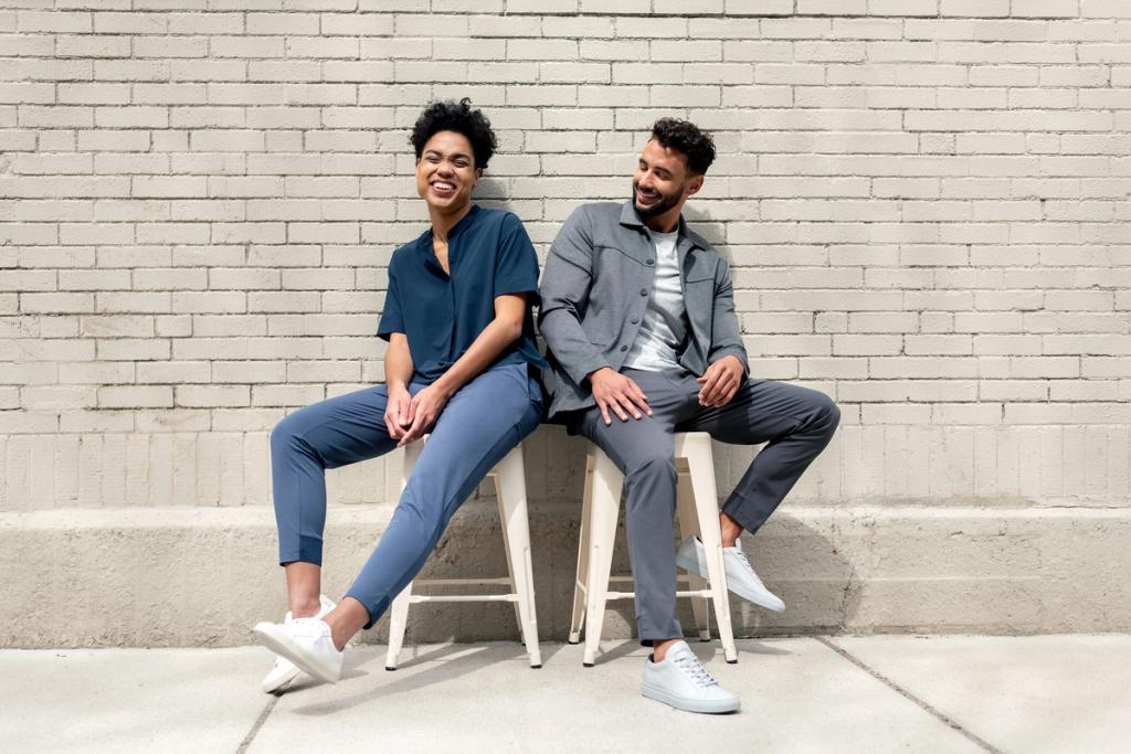Two people in business casual attire sitting on stools laughing against a gray brick wall