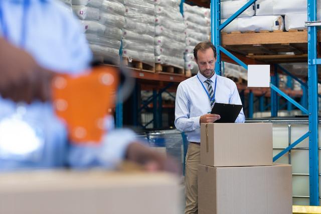 Warehouse manager in shirt and tie checking a clipboard beside stacked cardboard boxes, with racking and pallets behind