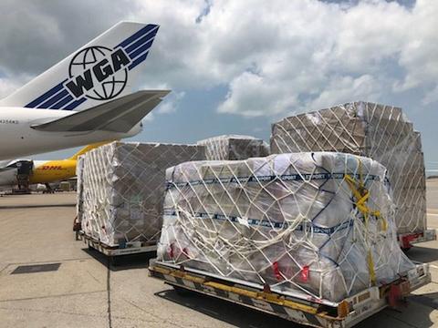 Netted cargo pallets staged on an airport tarmac beside a WGA cargo Boeing 747, with DHL aircraft in the background.