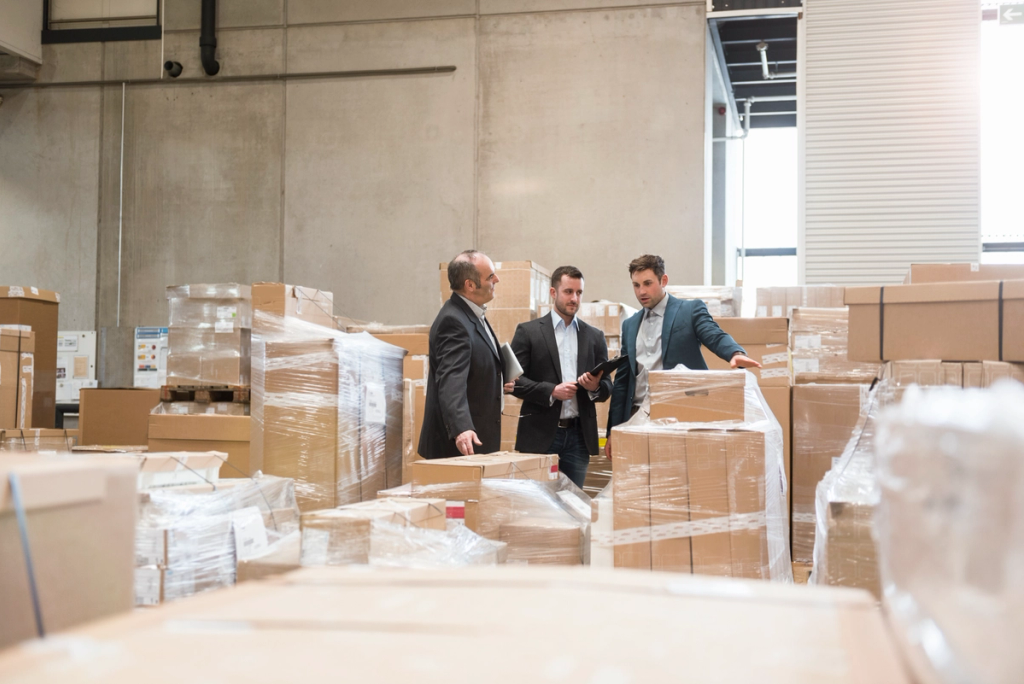 Three businessmen in suits talking among shrink-wrapped pallets and cardboard boxes in a warehouse