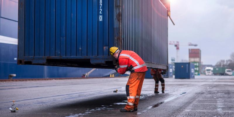 Worker in orange high-vis gear and yellow hardhat guiding a suspended blue shipping container at a yard