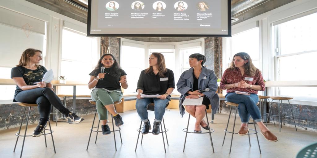 Five women on stools speak on a panel beneath a screen of speaker headshots in a bright loft