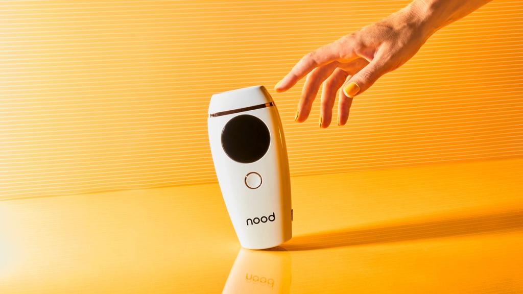 White Nood IPL hair removal device standing upright as a hand reaches toward it on orange
