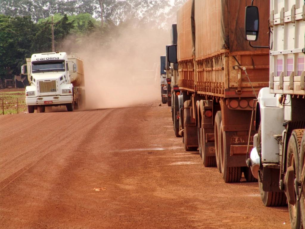 Line of cargo trucks parked on a red dirt road as a white tanker truck approaches, kicking up dust