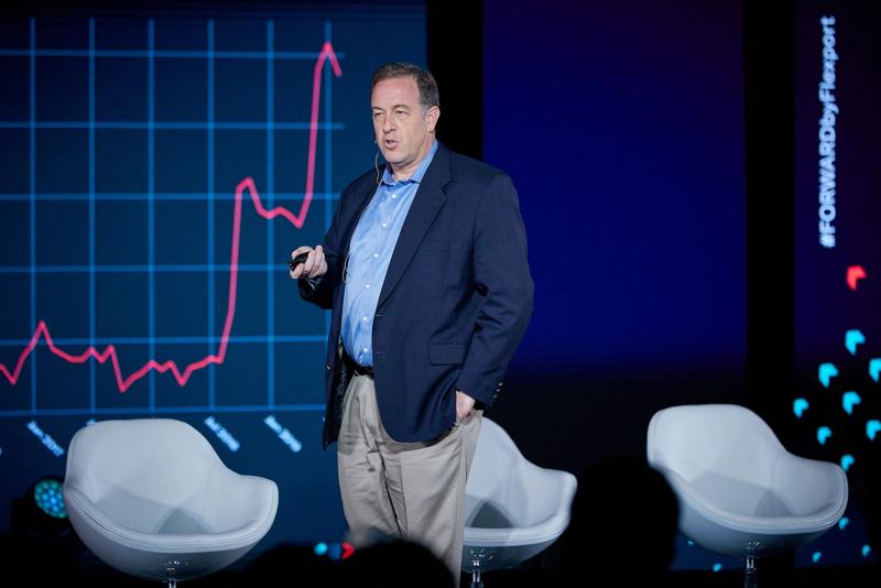 Man in a navy blazer presenting on stage in front of a large rising line graph at a Flexport FORWARD event