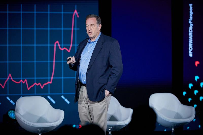 Man in a navy blazer presenting on stage in front of a large rising line graph at a Flexport FORWARD event