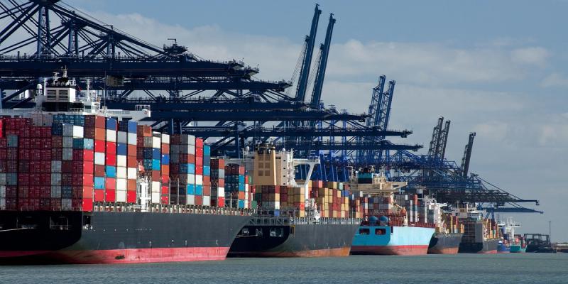 Row of container ships berthed under gantry cranes at a port, loaded with stacked containers