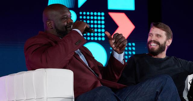 Two men laughing on stage, one in a maroon blazer speaking into a microphone, at a conference