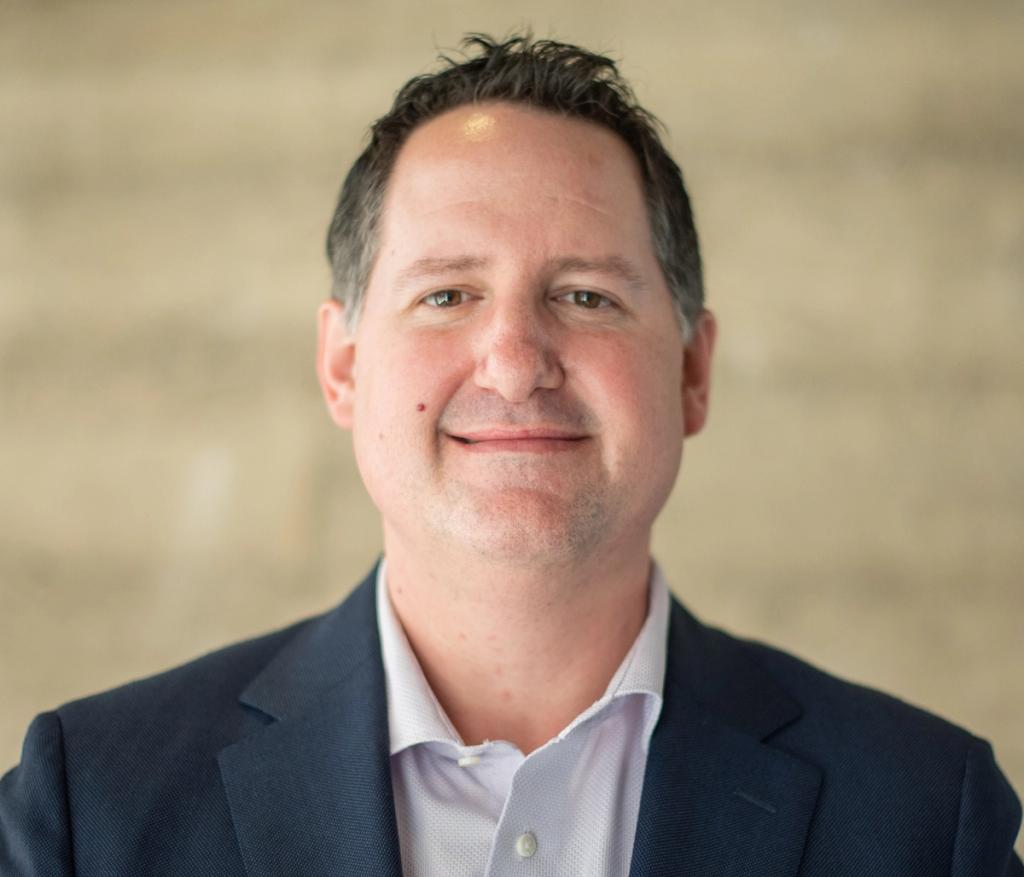 Headshot of a smiling man in a navy blazer and light dress shirt against a neutral background