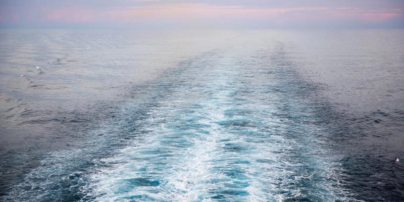 Foaming wake trailing from a moving ship across calm open ocean at dawn under a pink-tinged sky.
