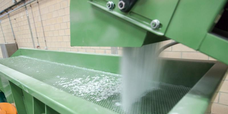 Green industrial recycling machine shredding white plastic flakes onto a conveyor in a tiled facility