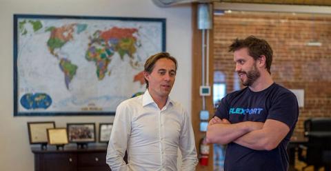 Two men talking in an office, one in a Flexport t-shirt, beside a framed world map on a brick wall