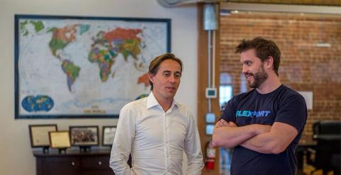 Two men talking in an office, one in a Flexport t-shirt, beside a framed world map on a brick wall