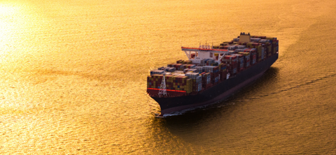 Aerial photo of a fully loaded container ship sailing on open water lit gold by a low sun.