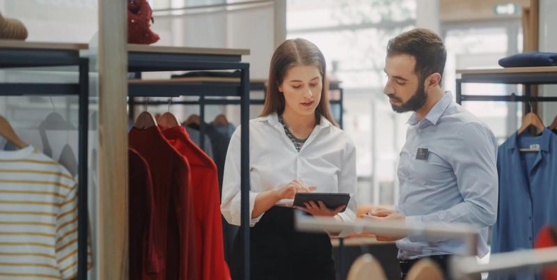 Two retail staff in white and blue shirts review a tablet beside racks of clothing in an apparel store