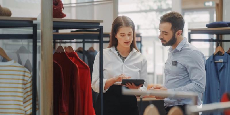 Two retail staff in white and blue shirts review a tablet beside racks of clothing in an apparel store