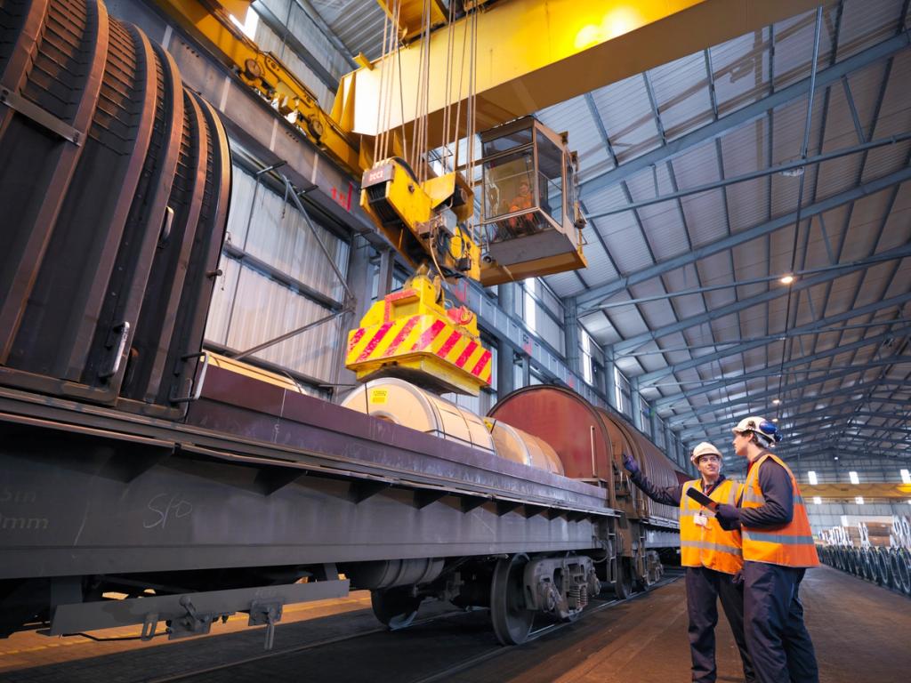 Workers in hi-vis vests and hard hats inspect steel coils on a rail wagon as an overhead crane with a clamp lifts cargo in a warehouse