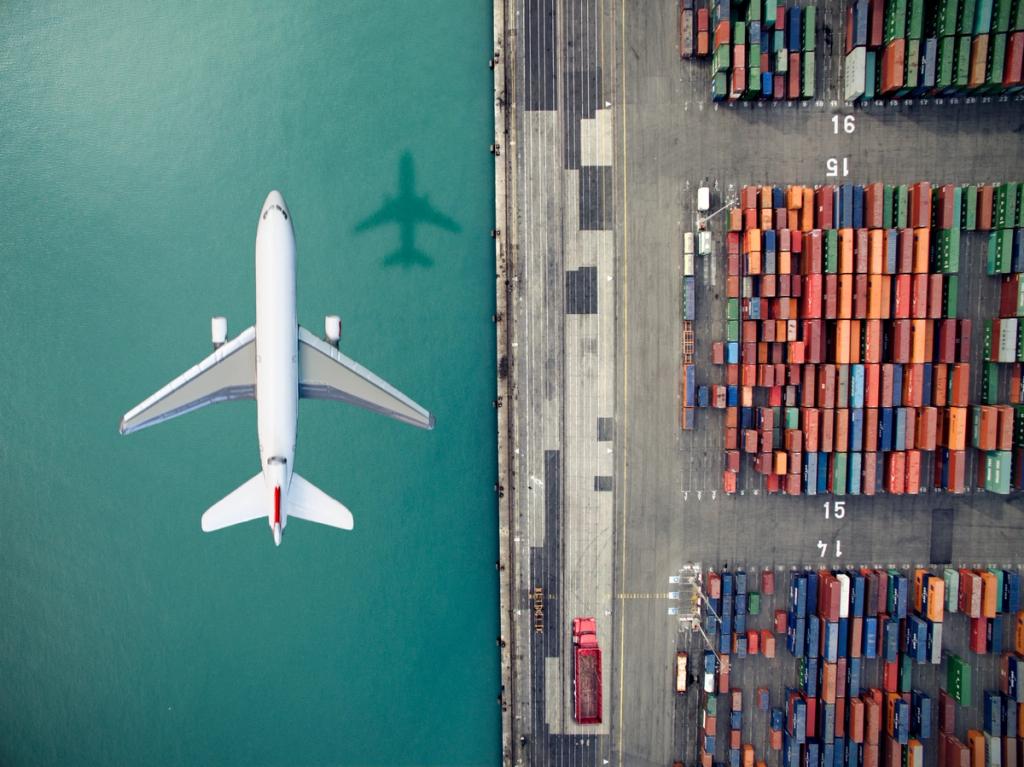 Aerial view of a passenger jet flying over teal water beside a container yard with rows of stacked shipping containers