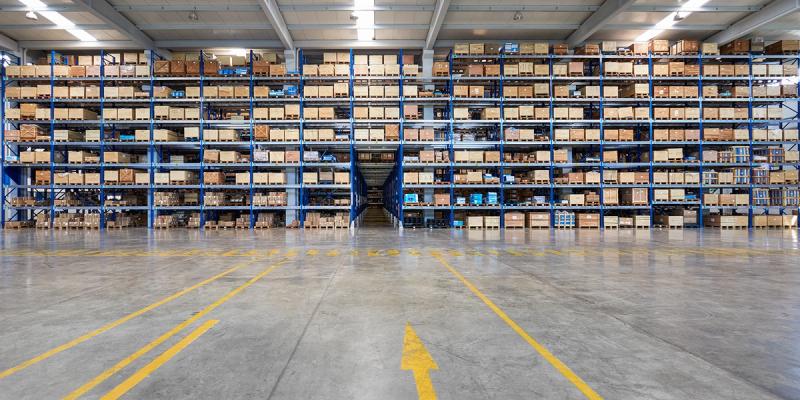 Vast warehouse with tall blue racks of stacked cardboard boxes and yellow floor arrow markings