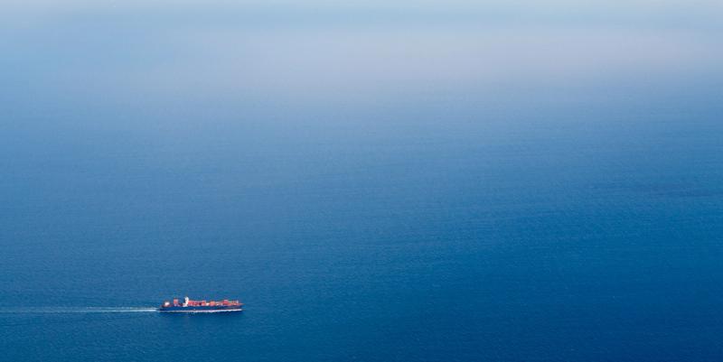 A lone red container ship crosses a vast expanse of deep blue open ocean.
