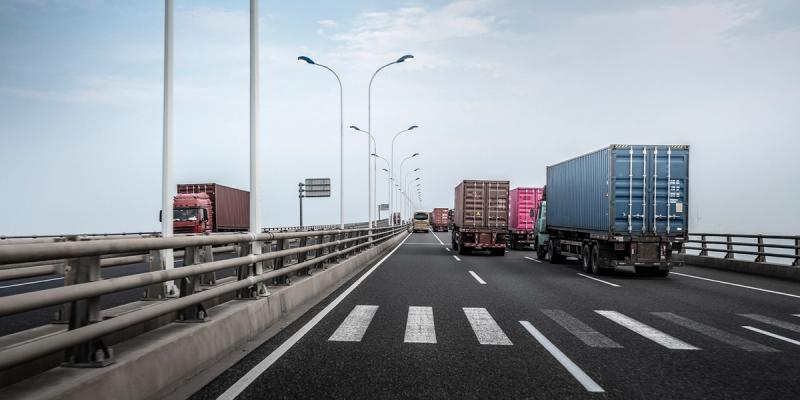 Container trucks driving across a multi-lane highway bridge lined with lampposts under a hazy sky