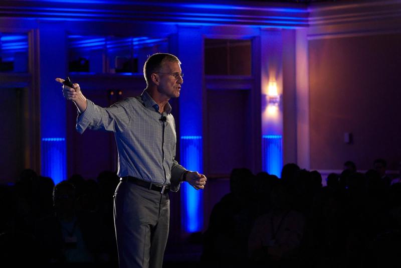 Man in checked shirt gestures while presenting on a stage lit with blue lighting before an audience.