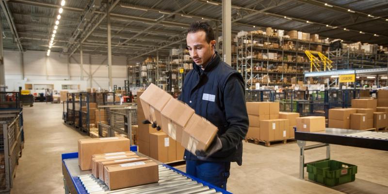 Warehouse worker in a vest stacking sealed cardboard boxes on a roller conveyor amid storage racks